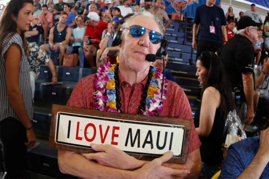 Bill Walton posing with a sign that reads, "I love Maui" with a headset on in crowded bleachers.