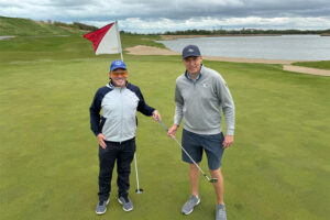 Two men posing on a golf course in front of the hole.