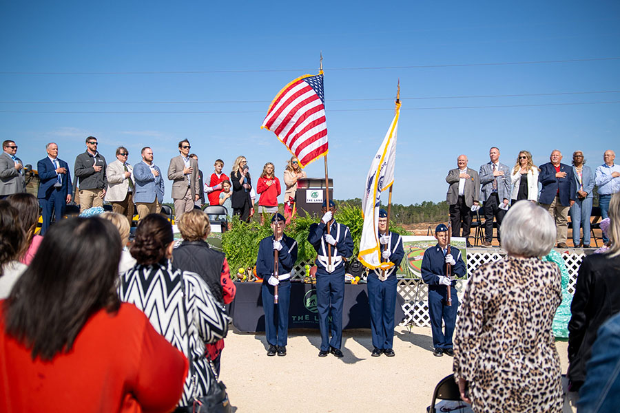 Group of people standing for the national anthem at the Saraland Groundbreaking ceremony. 4 military officials stand with the American flag and state flag.